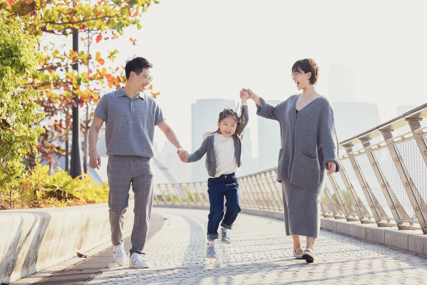 Bonnie, Vincent and Emma walking hand-in-hand along a sunny path at West Kowloon, Emma jumping playfully between her parents
