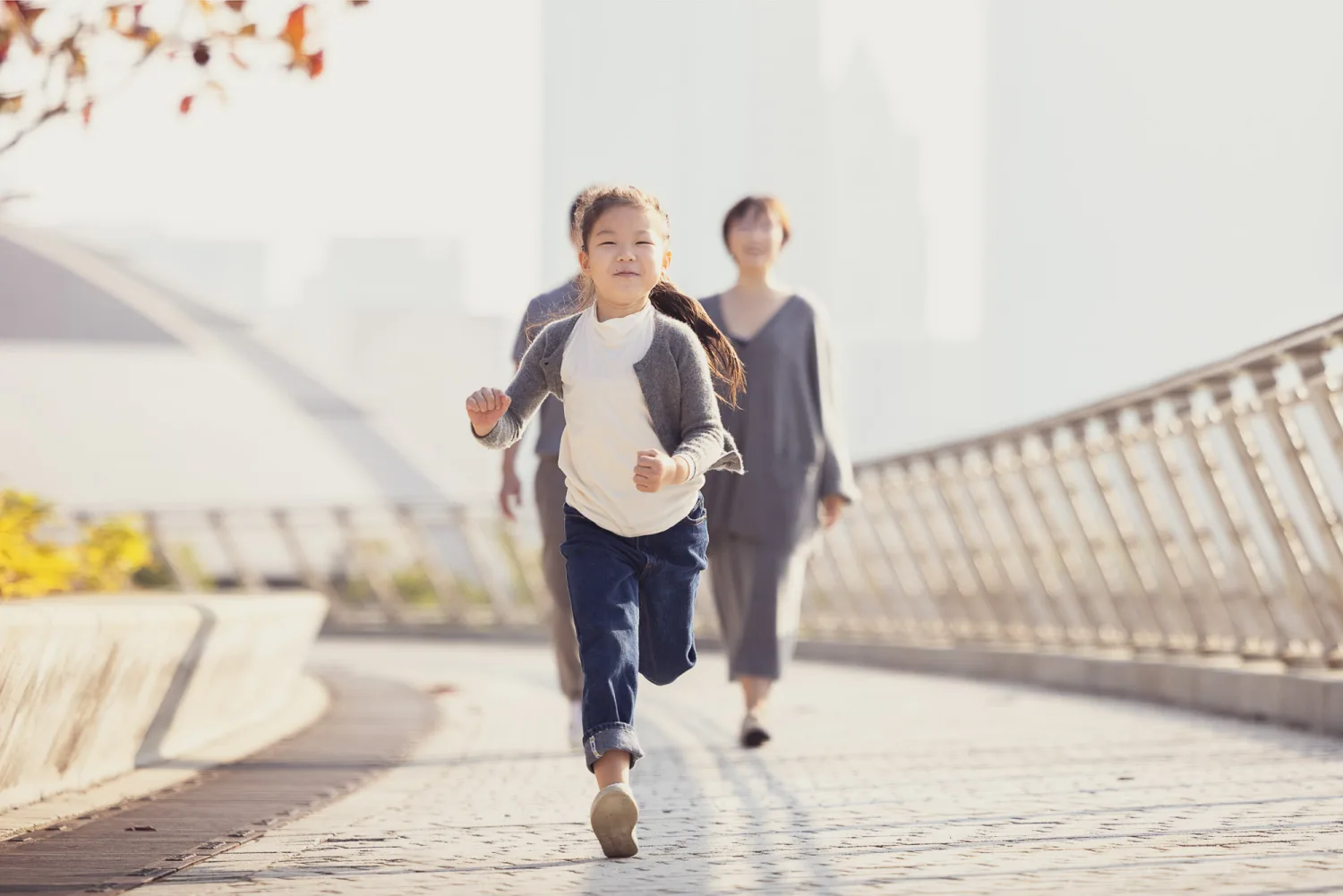 Emma running towards the camera at West Kowloon, smiling brightly in her white top and grey cardigan while Bonnie and Vincent follow behind