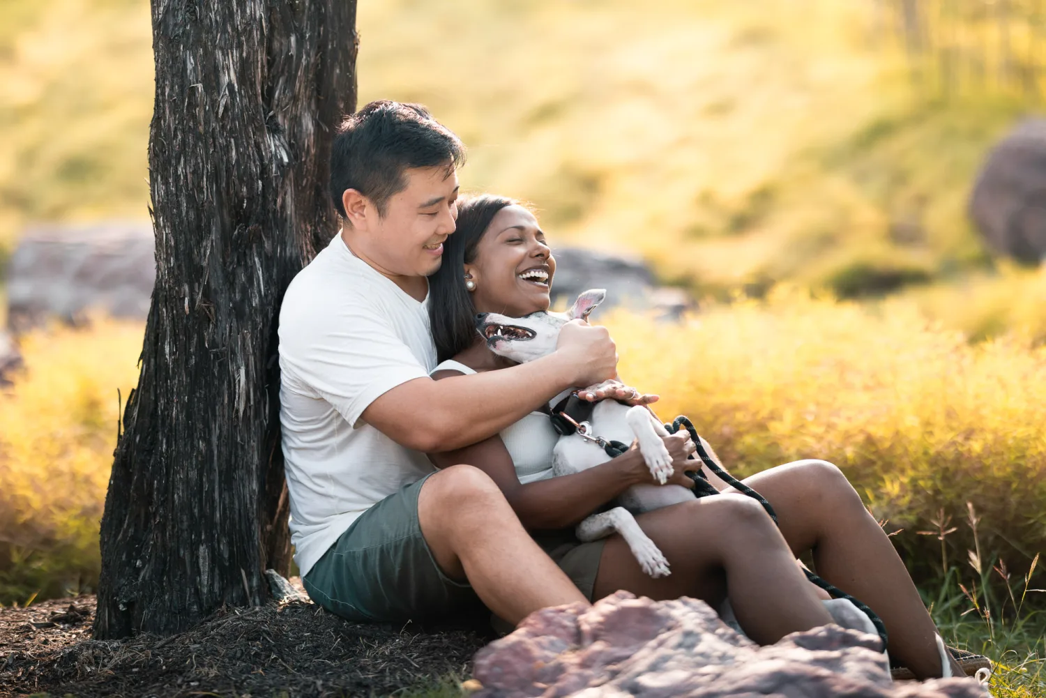 Dan and Jessica leaning against a tree, laughing, with Luna in Jessica's lap